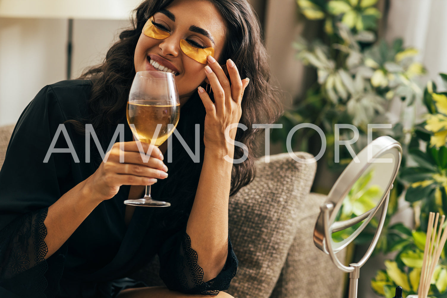 Smiling woman in satin robe relaxing at home. Beautiful female sitting on a couch holding a wineglass adjusting eye patch.	