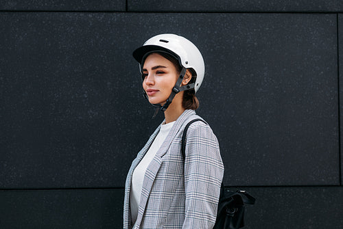 Businesswoman in white cycling helmet wearing backpack standing against a black wall outdoors