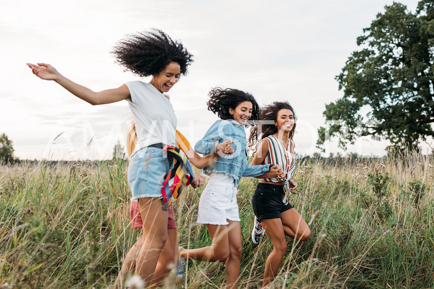 Four women holding hands running on field. Laughing friends having fun outdoors in summer.