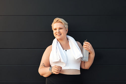 Smiling plus size woman sportswear standing at black wall. Healthy woman relaxing after training holding a bottle of water looking away.