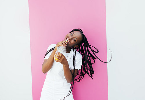 Cheerful woman with braids drinking a juice and looking at camera while standing at pink wall