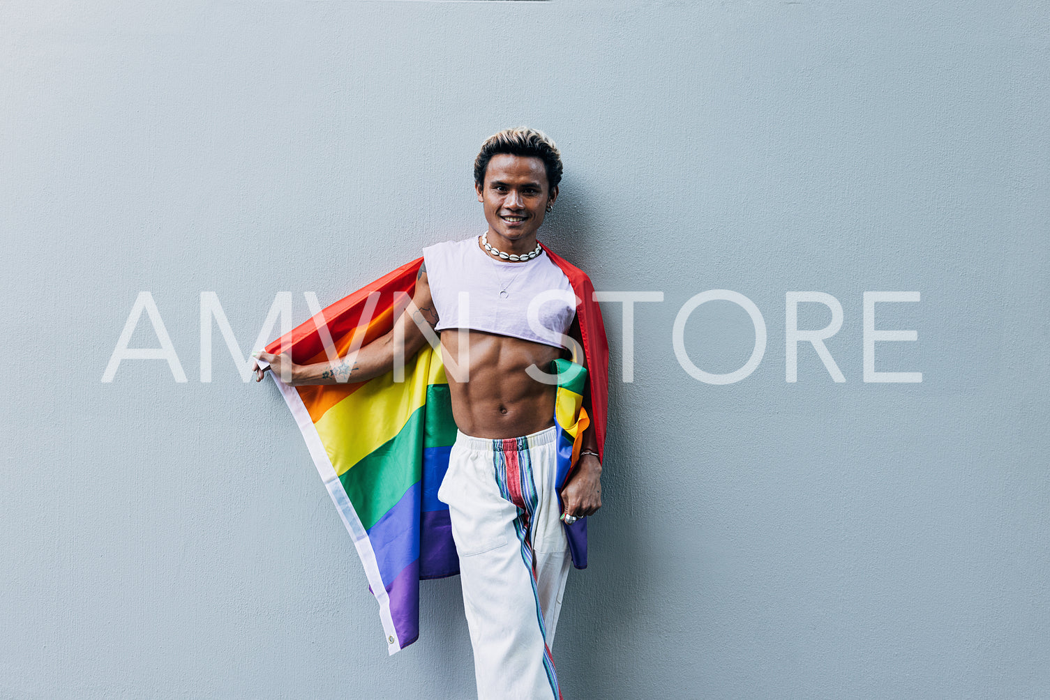 Happy guy with rainbow flag standing outdoors at grey wall and looking at camera wearing stylish clothes