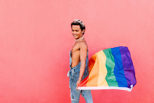 Side view of a young stylish guy walking with rainbow flag at pink wall