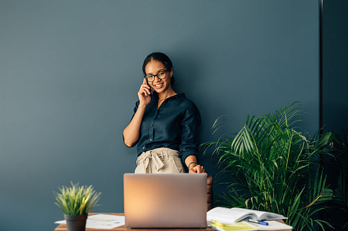 Smiling young woman standing at the table and looking on laptop screen while talking on cell phone