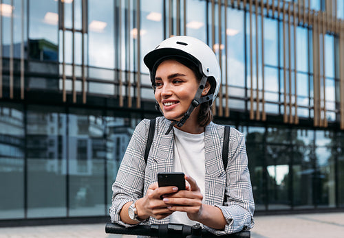 Smiling woman leaning on electric push scooter using a mobile phone while standing near an office building