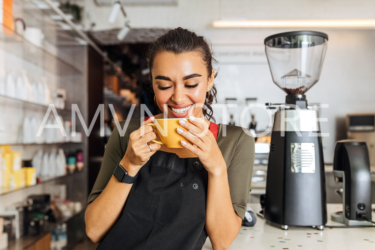 Happy barista standing inside coffee shop with closed eyes and holding a mug. Cheerful woman in apron standing at cafe counter.