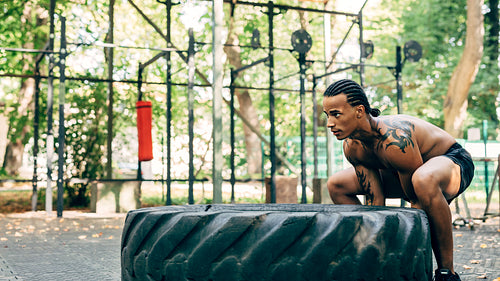 Side view of a bare-chested athlete doing squats with huge tire on sports ground outdoors