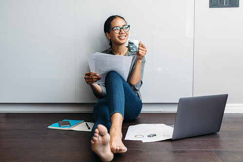 Smiling woman with a coffee mug holding documents. Young entrepreneur sitting on the kitchen floor looking away.