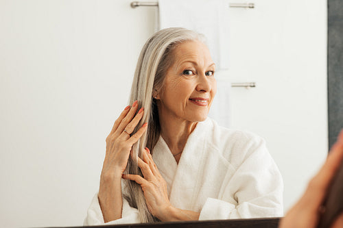 Woman with grey hair in the bathroom in front of a mirror. Senior female adjusting long hair with hands.