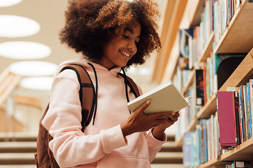 Smiling girl holding a book in library while standing at bookshelf