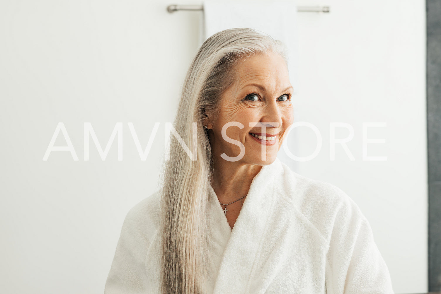 Smiling woman with long white hair at morning in bathroom. Aged female in bathrobe looking at her reflection in the mirror.