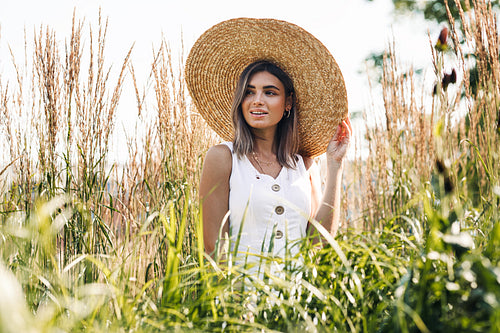 Young happy woman in big straw hat standing on the field and looking away. Stylish female walking outdoors.