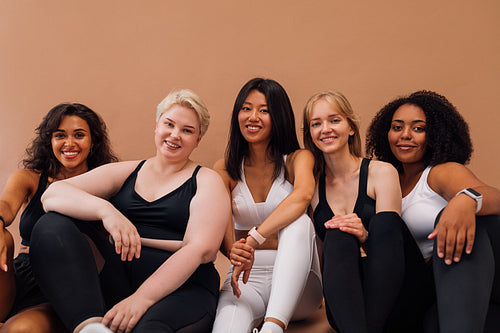 Five smiling women in fitness clothes sitting together on pastel background. Females with different body types relaxing after workout.