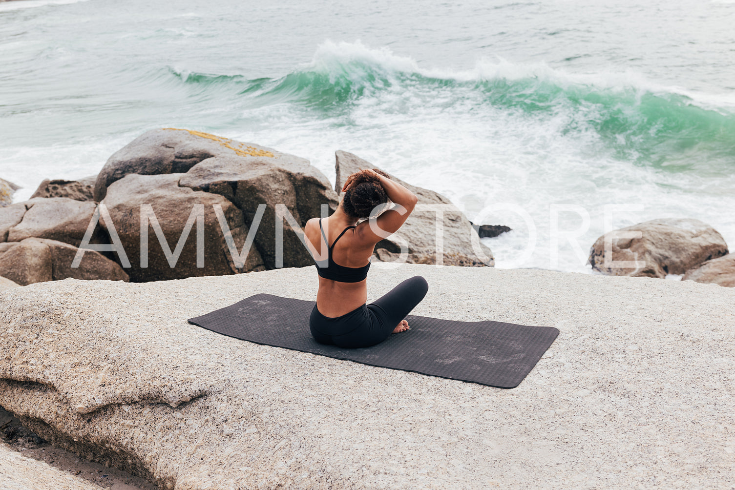 Rear view of young woman sitting on mat and warming up her neck by ocean. Female doing yoga and looking on waves.