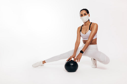 Mixed race woman with face mask sitting in a studio with a medicine ball