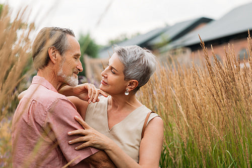 Two aged people standing together in the field. Senior wife and husband are looking at each other and embracing the outdoors.