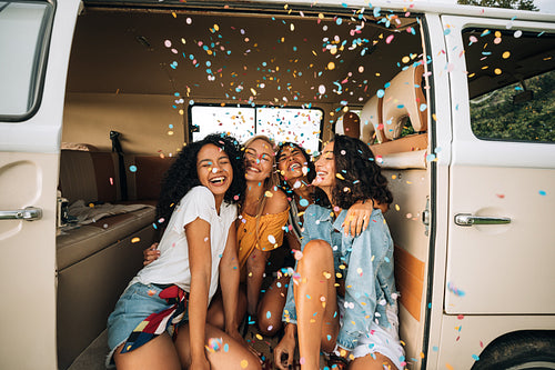 Group of happy women sitting in a camper van door under confetti. Friends having fun on a road trip.