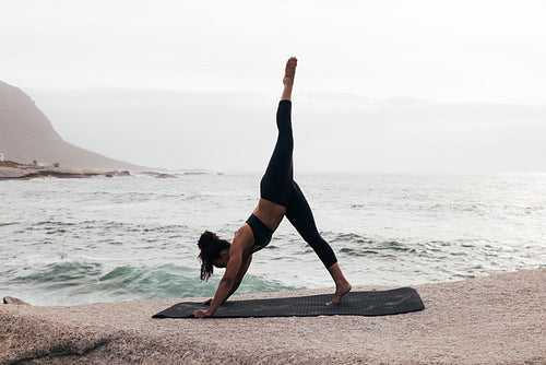 Side view of young woman doing Three-Legged Down Dog pose by ocean at sunset