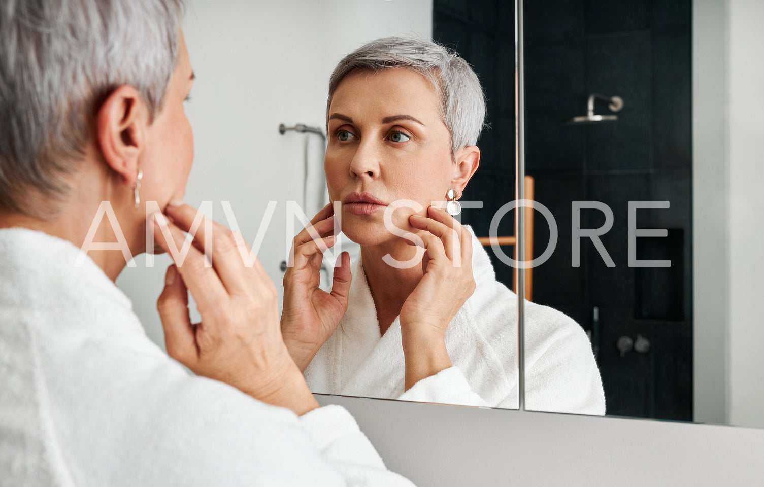 Mature woman with short grey hair touching her face in front of a bathroom mirror