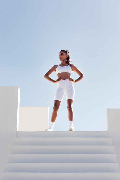 Full length of a slim confident woman looking away while standing on the top of stairs. Sportswoman in white fitness attire against skies.