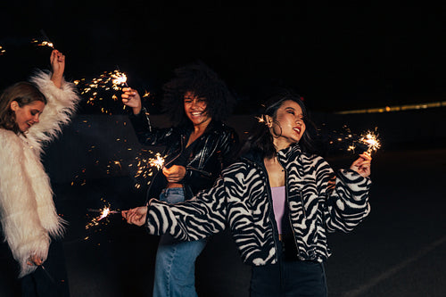 Laughing female girlfriends dancing outdoors with sparklers at night