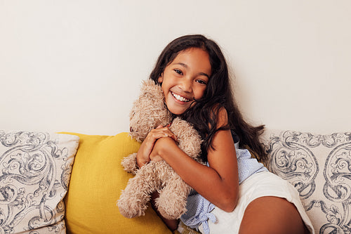 Girl with toy on the sofa. Smiling kid hugging a teddy bear and looking at camera.