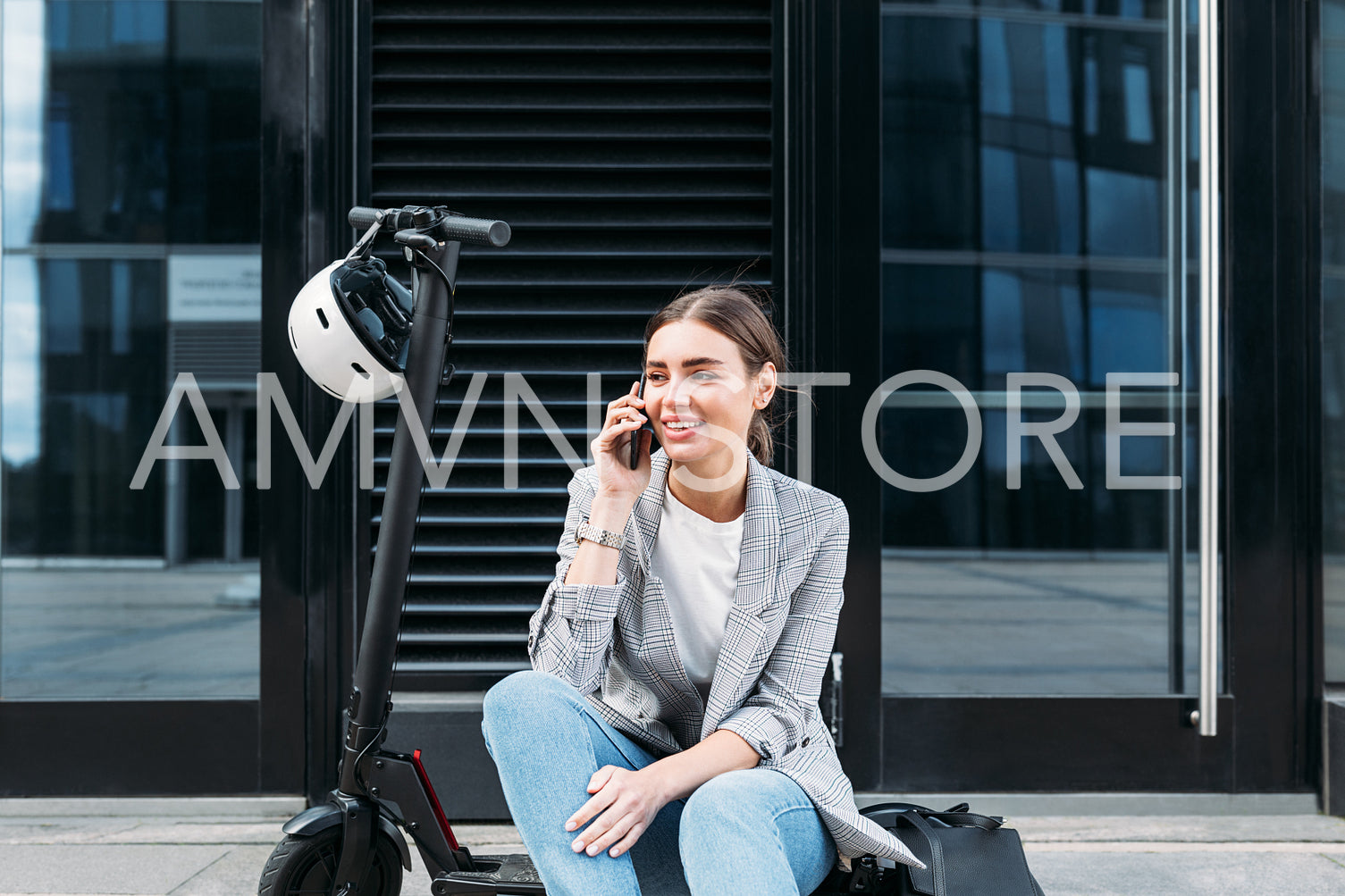 Young businesswoman sitting on an electric push scooter near a building and talking on mobile phone