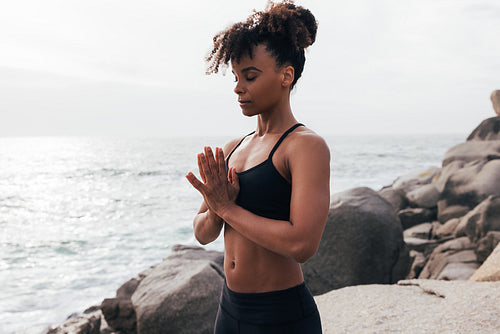 Young woman with folded hands standing at sunset by ocean. Female meditating outdoors with closed eyes.