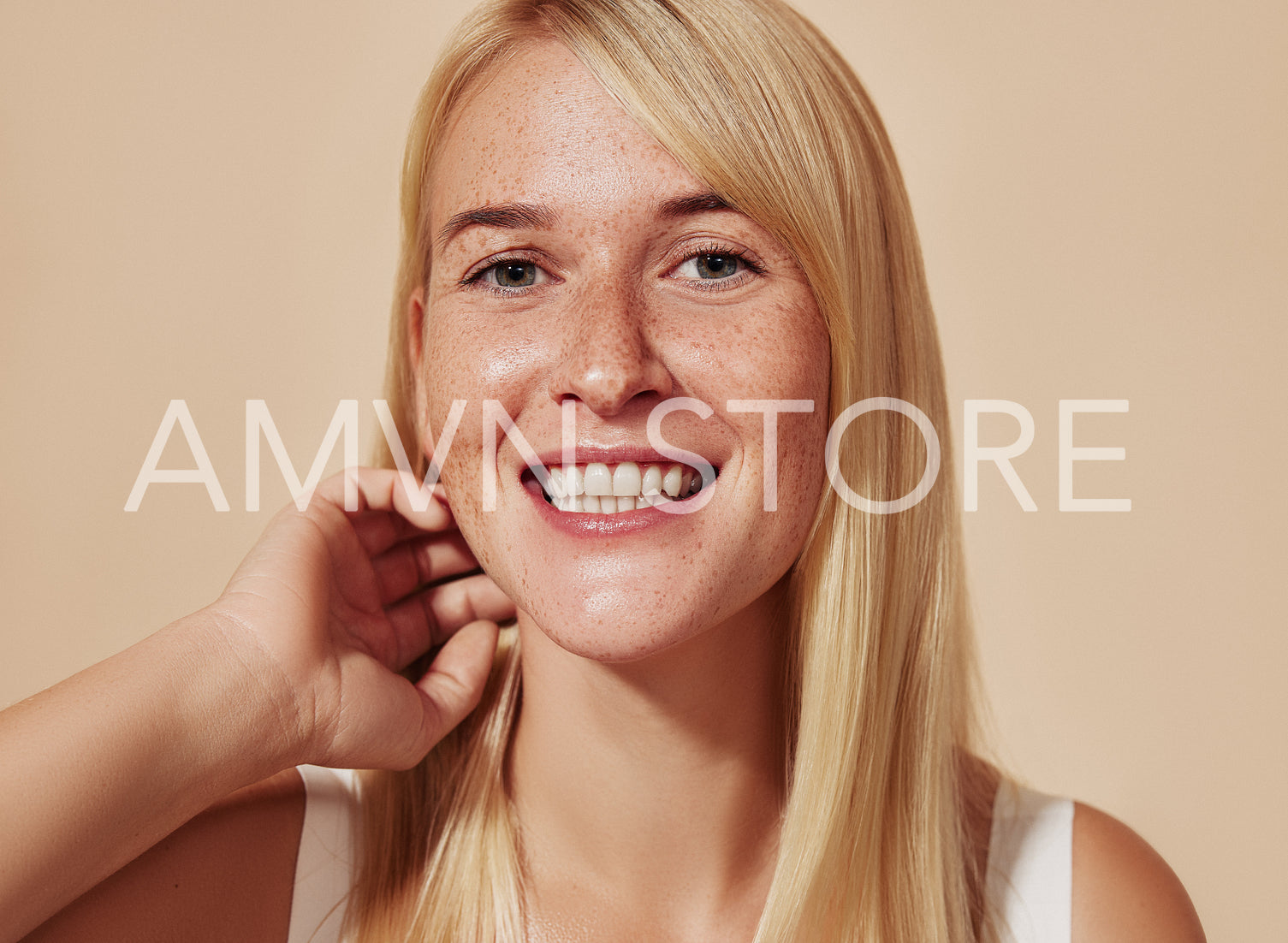 Young happy female with blond hair looking at camera in a studio. Beautiful smiling woman with freckles adjusting her hair over a beige backdrop.