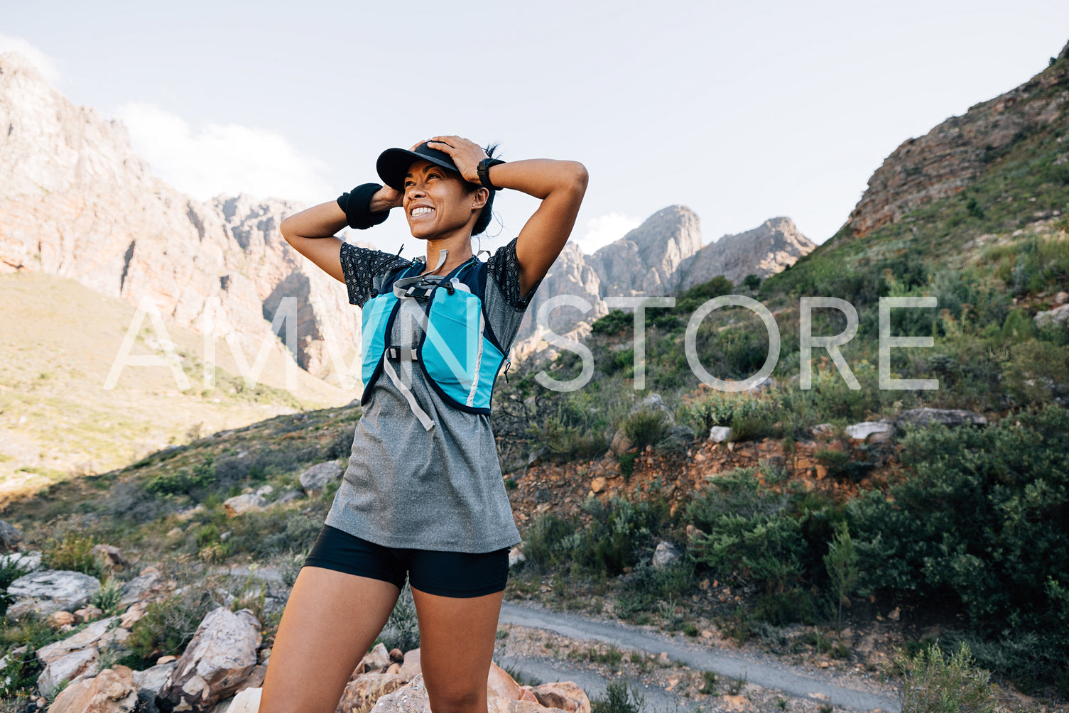 Happy woman hiker with hands on cap. Smiling female enjoying the view while standing in valley and looking at the mountains.