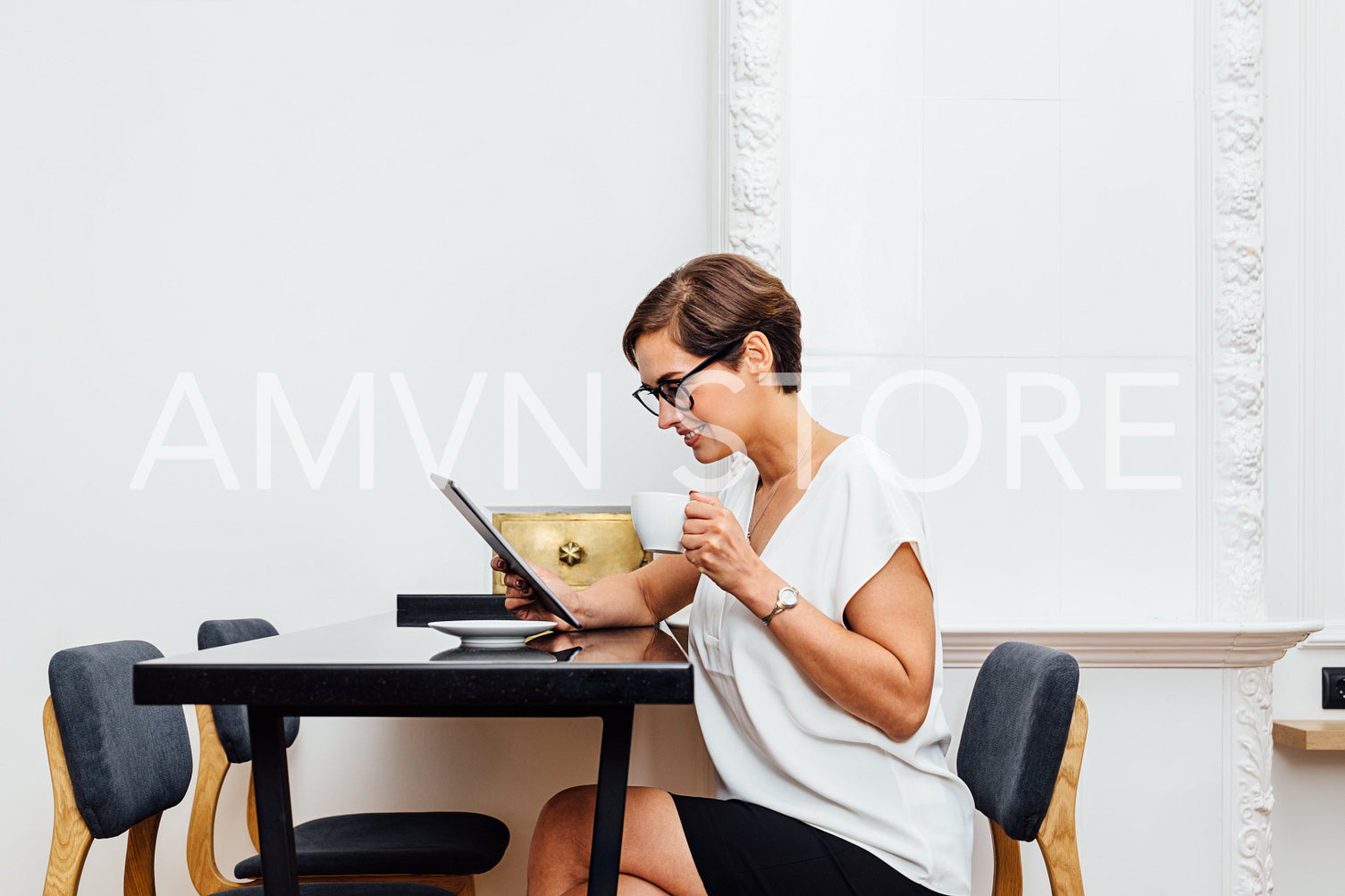 Side view of a woman sitting at table and reading from digital tablet at apartment. Businesswoman holding a cup and looking on tablet screen.	