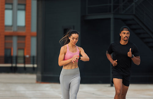 Two athletes sprinting on a rooftop. Young woman checking fitness tracker while running.