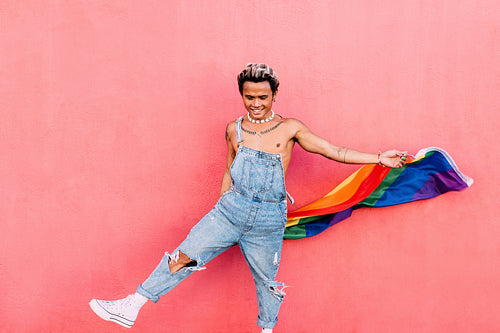 Smiling guy dancing with rainbow LGBT flag against pink wall outdoors