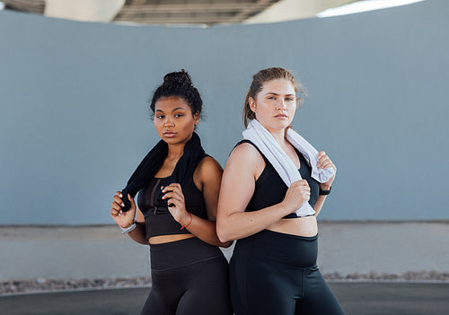 Two plus-size women in fitness wear standing back to back looking at camera. Two young females with towels posing after a workout.