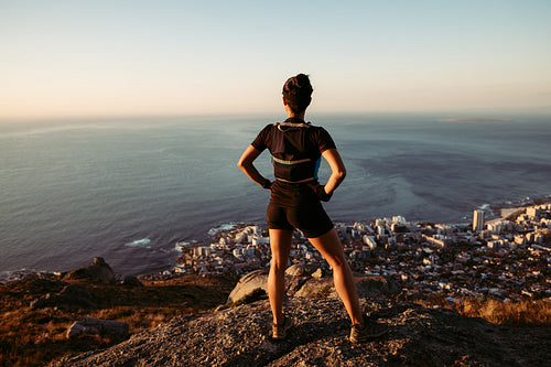 Rear view of sportswoman enjoying the view from the top. Female runner in sportswear taking a break on a hill at sunset.