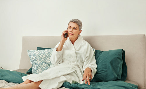 Senior woman with short grey hair talking on a mobile phone from hotel room lying on a bed