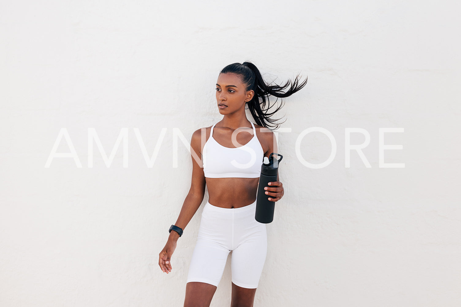 Portrait of a slim sportswoman with a shaker at a white wall. Female in white fitness attire holding a bottle.