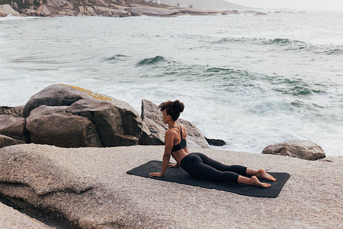 Female in cobra yoga pose at seaside. Woman in sportswear doing stretching workout on mat by ocean.