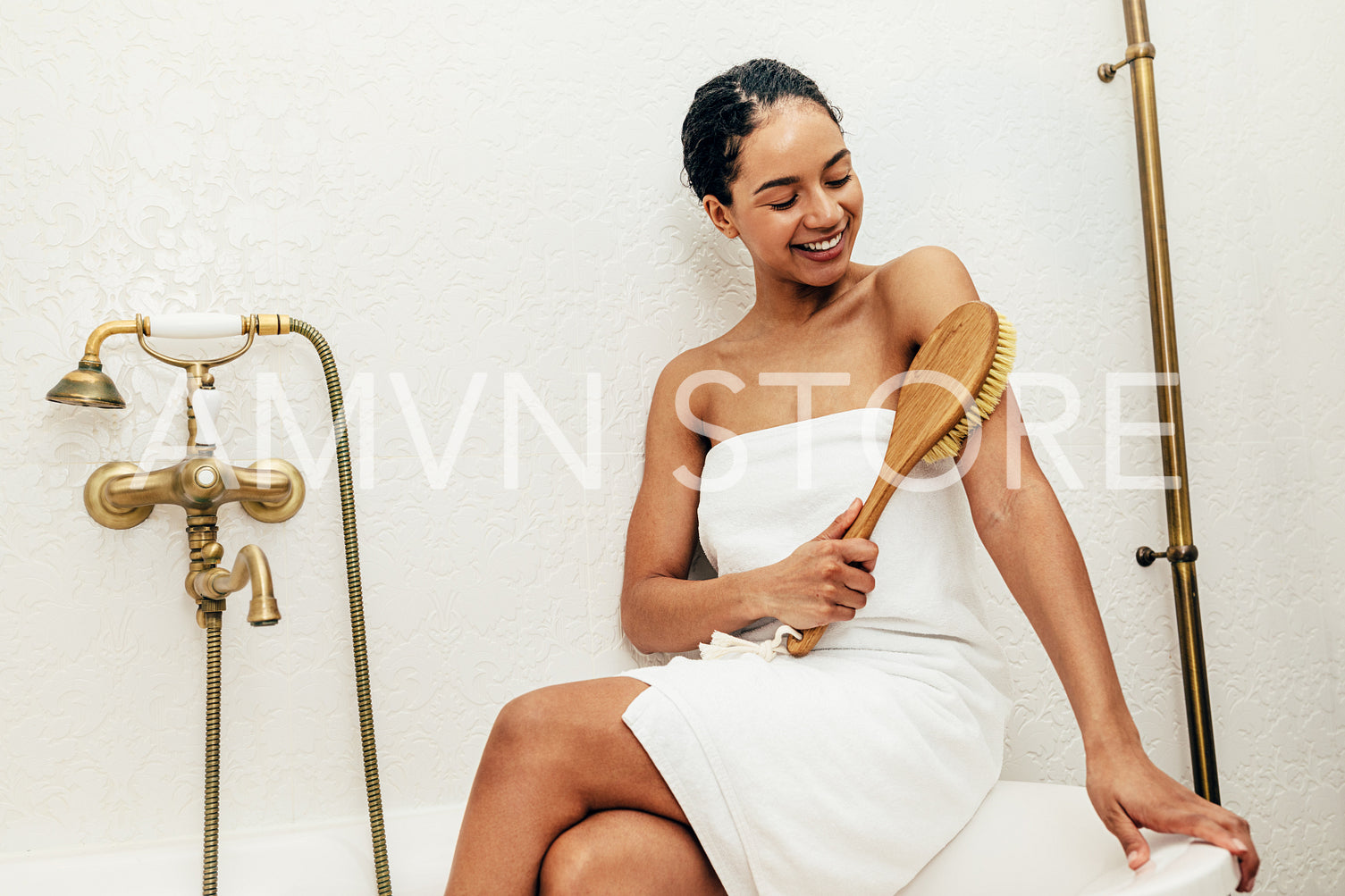 Smiling woman wrapped in white towel sitting on bath and dry brushing her arm	