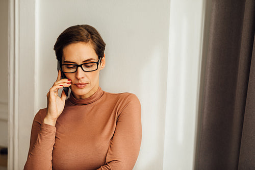 Relaxed businesswoman in casuals standing at wall and talking over mobile phone in hotel room