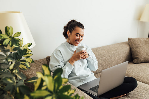 Young smiling woman holding a cup while sitting on a sofa with laptop on legs