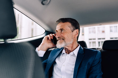 Smiling businessman talking on a cell phone while sitting on a backseat of a taxi