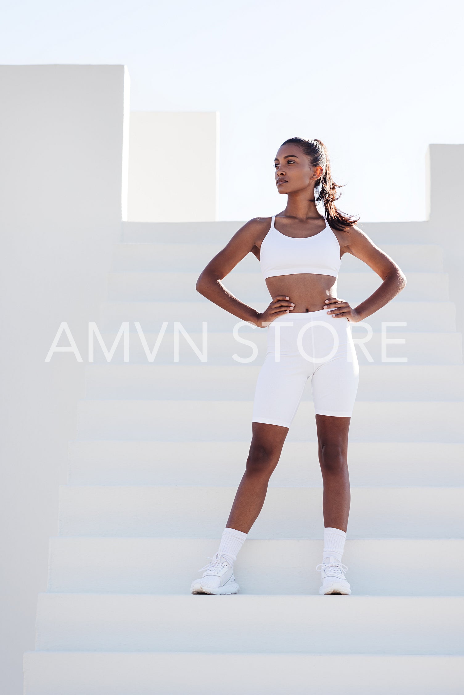 Slim and healthy woman in white fitness attire relaxing on stairs. Full length of a young female with hands on hips looking away outdoors.