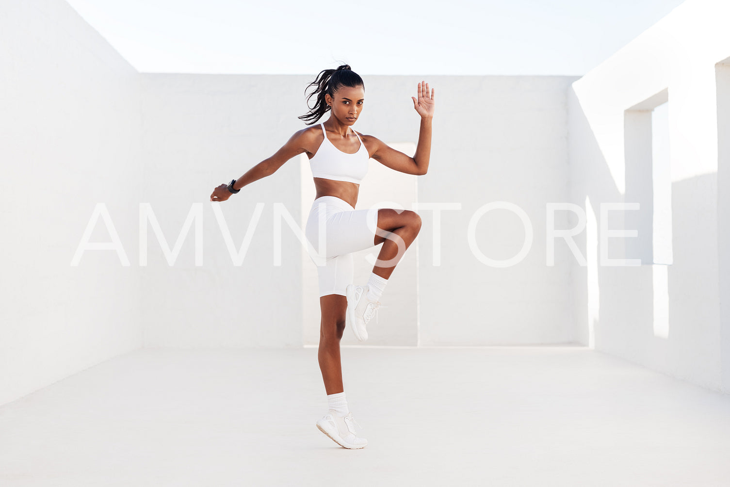 Young woman athlete jumping in white outdoor studio. Slim female doing cardio exercises. Sportswoman practicing high-intensity exercises.