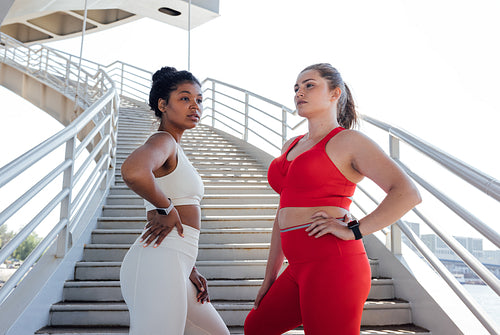 Two confident plus-size women in fitness attire stand together on a staircase outdoors. Females with the same body type posing outdoors after fitness training.