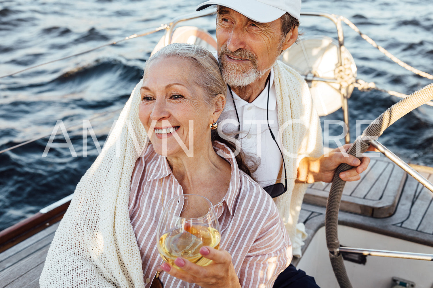 Elderly couple sitting together on a private yacht and wrapped in a plaid. Smiling caucasian woman drinking a wine while her husband stearing a wheel.	