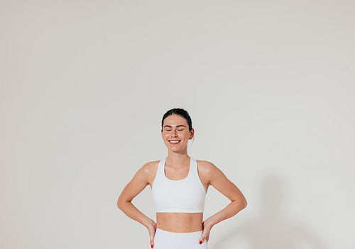 Smiling woman with closed eyes and hands on hips wearing white fitness attire standing at white wall