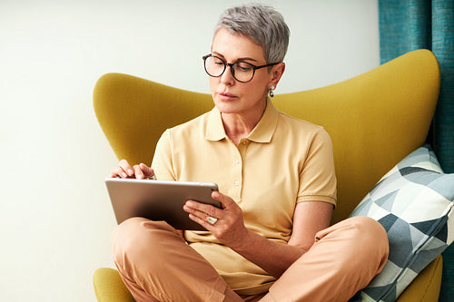 Mature woman sitting with crossed legs on armchair using a digital tablet