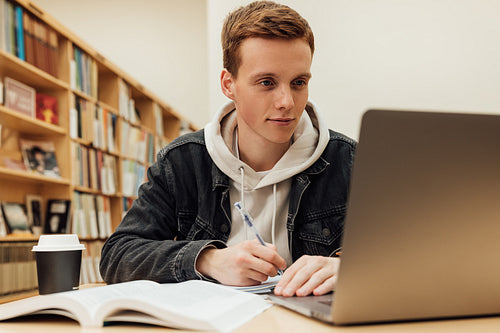 Male student writing and looking on laptop. Guy in casuals preparing exams at desk in library.