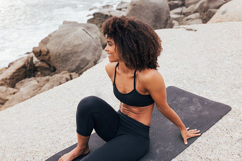 Smiling woman sitting on a rock near an ocean. Cheerful female relaxing on yoga mat looking at ocean.